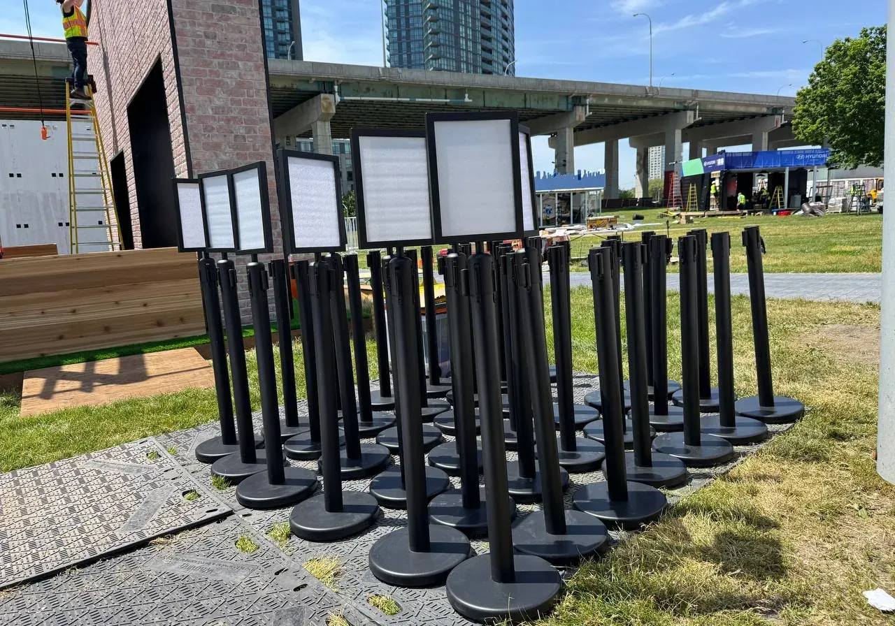 Rows of black belt stanchions fitted with empty A4 sign frames, staged on grass before an event opens