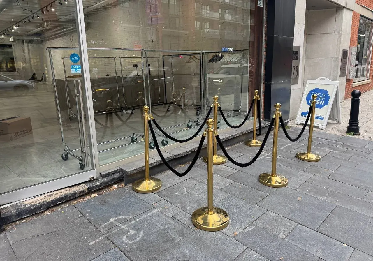 Six gold ball-top stanchions with black velvet ropes forming a short queue on a stone sidewalk outside a glass shop front
