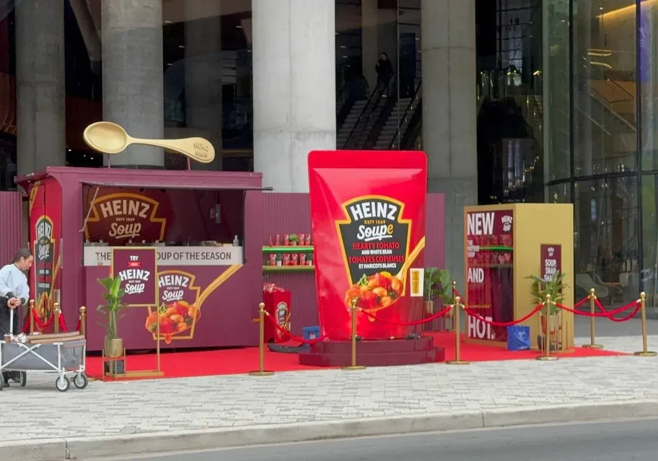 Gold ball-top stanchions and red velvet ropes fencing off a red carpet promotional booth on a plaza outside a glass office tower